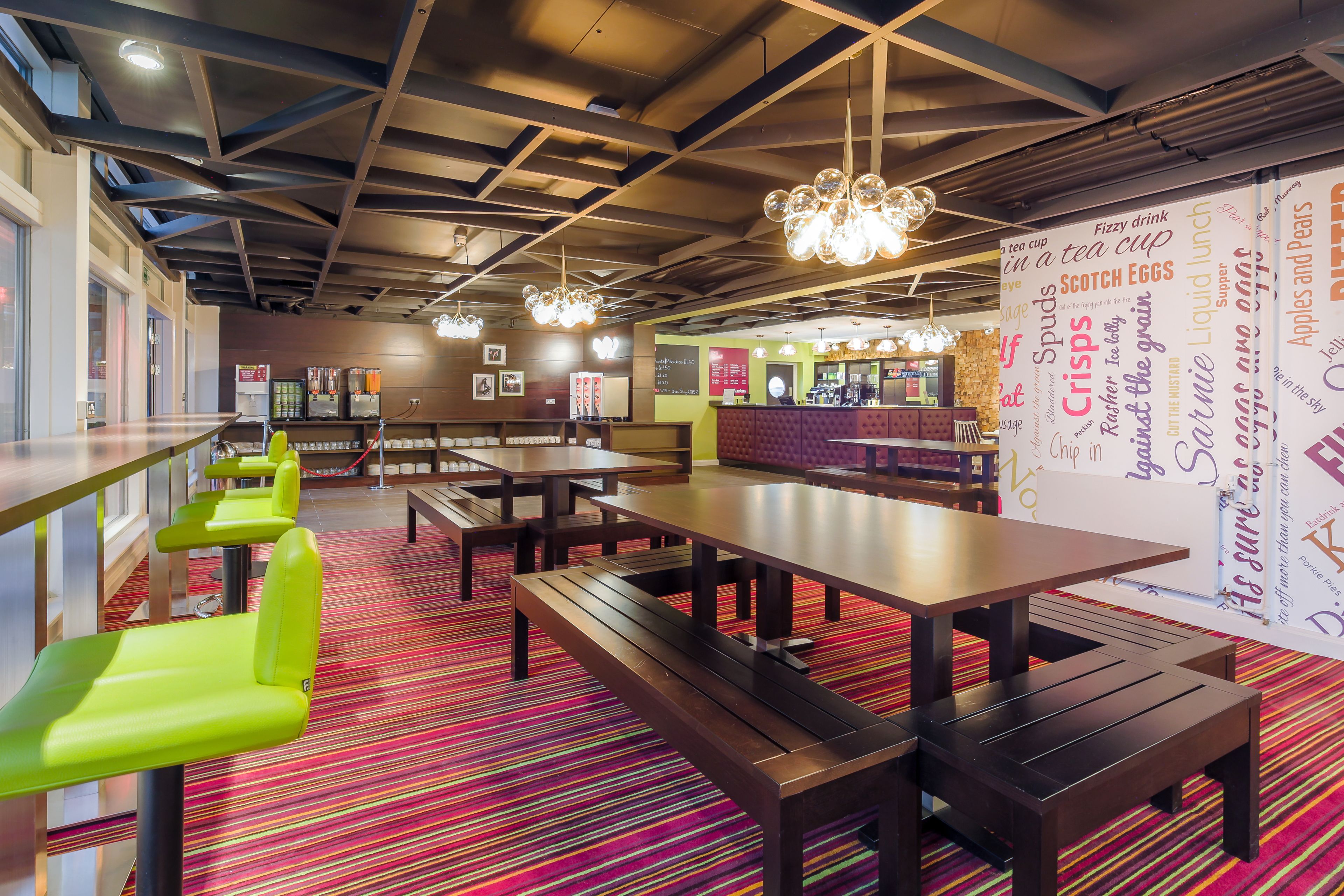 A modern restaurant interior with wooden tables and benches, a colorful striped carpet, and a menu board in the background.