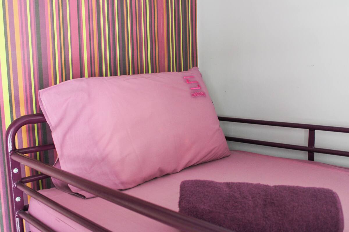 A single bed with a pink mattress and a purple blanket, a metal frame, and a striped wall in the background.
