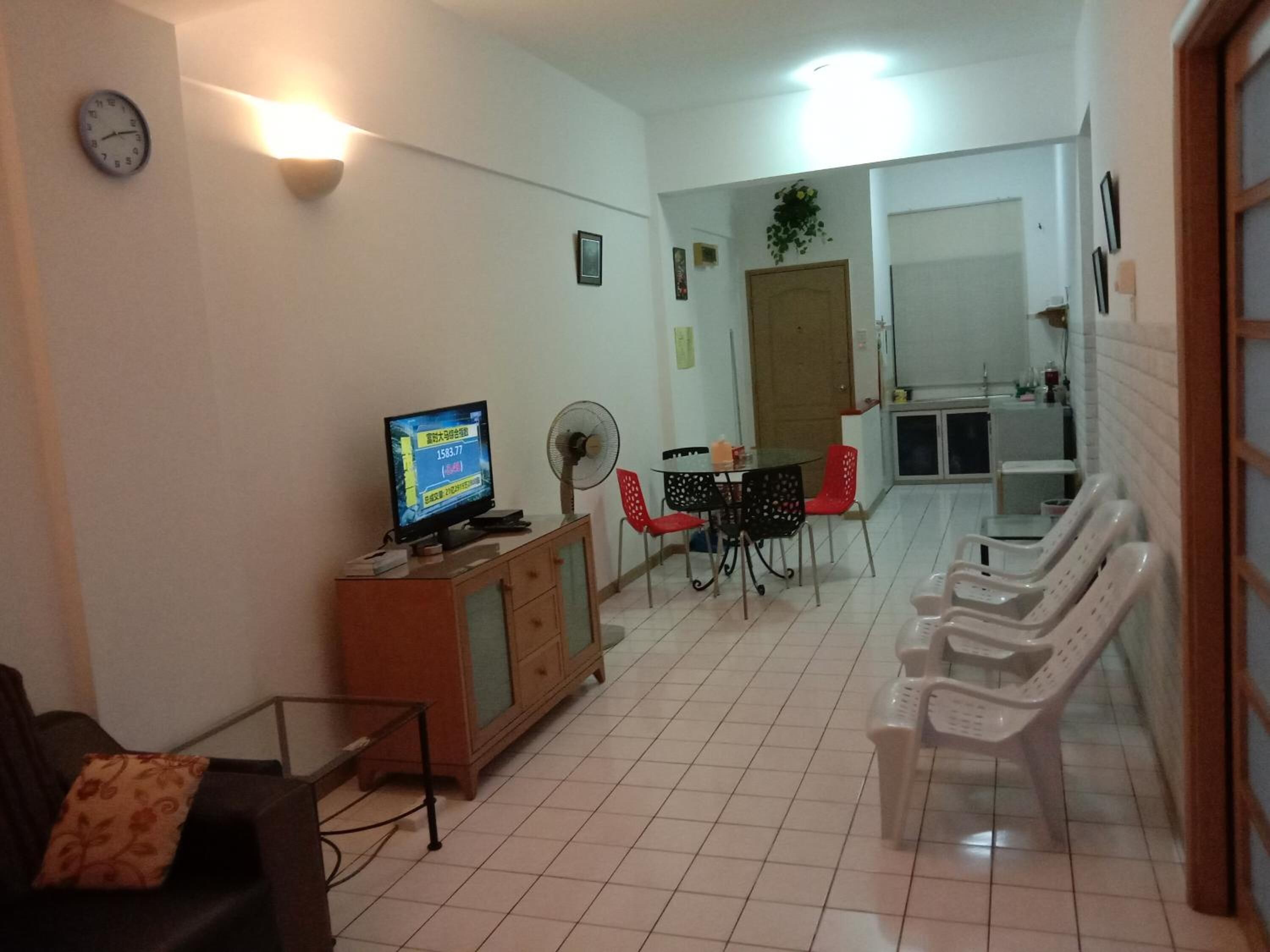 A living room with a TV, a fan, a dining table with chairs, and a kitchen area in the background.