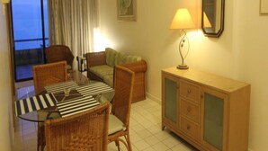 A room with a dining table, chairs, a sofa, a cabinet, a lamp, and a large window with curtains. - Paradise Lagoon Apartment (Port Dickson)
