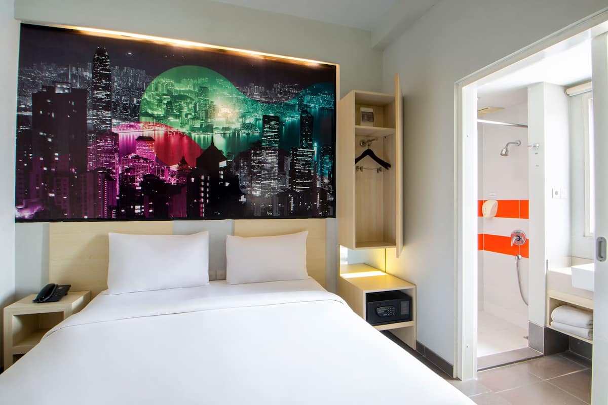 A hotel room with a bed, a nightstand with a phone, a wall-mounted TV, a bathroom with a shower, and a cityscape artwork on the wall.