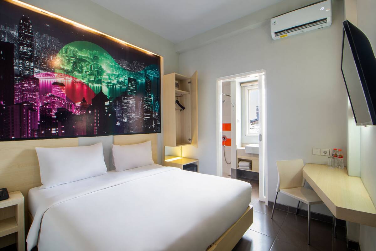 A hotel room with a large bed, a desk, a chair, a wardrobe, and an air conditioning unit.