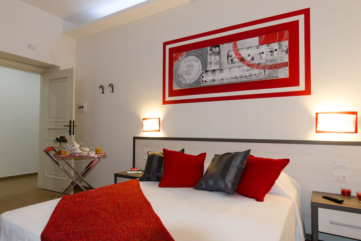 A hotel room with a bed, a table with a red blanket, a wall-mounted painting, and bedside lamps.