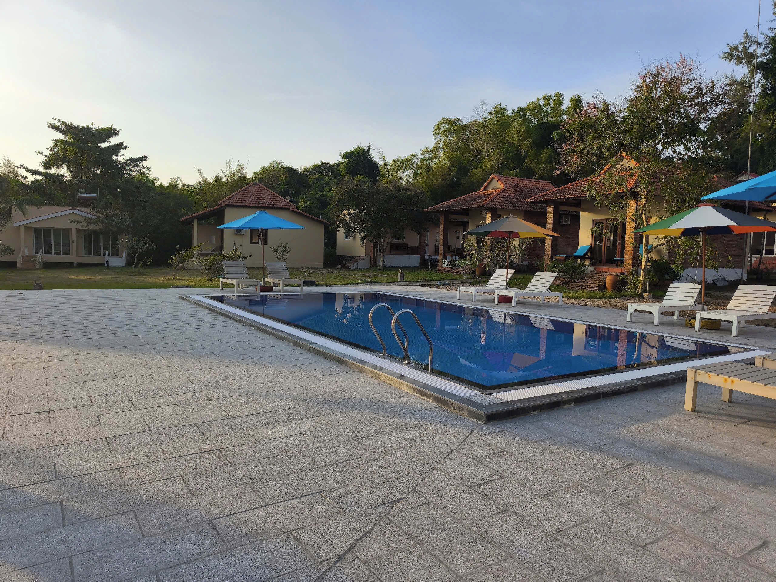 Ein rechteckiger Pool mit einer Metallleiter, umgeben von einer gepflasterten Fläche mit Liegestühlen und Sonnenschirmen.