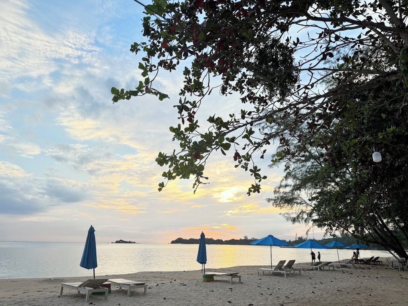 Ein Strand mit Liegestühlen und Sonnenschirmen, ein ruhiges Meer und ein Sonnenuntergang im Hintergrund.