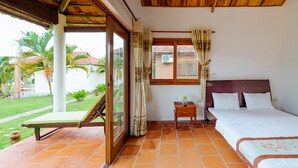 A bedroom with a bed, a nightstand, a window with curtains, and a view of a garden. - Vung Bau Resort (Phu Quoc)