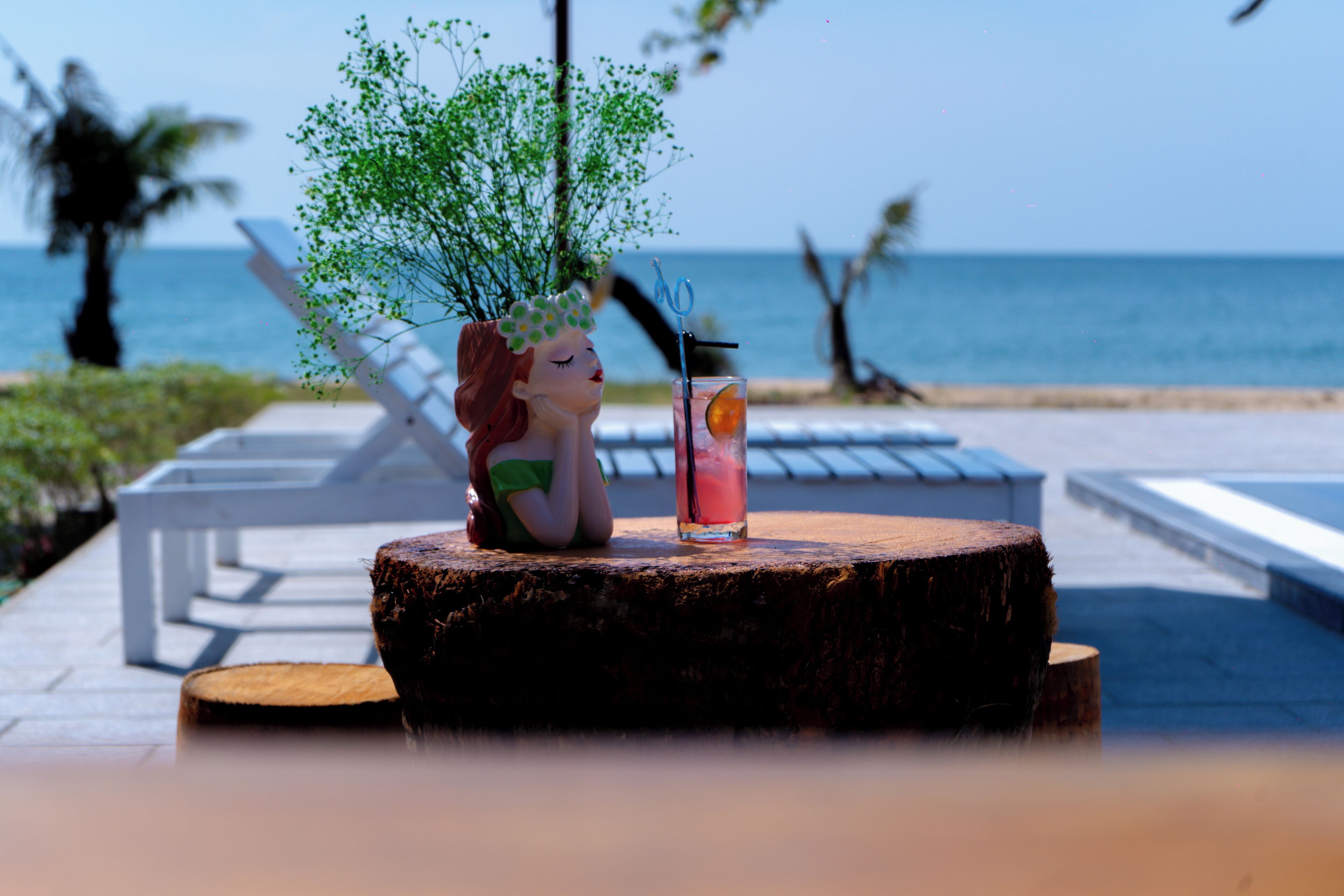 Una estatua de una mujer sentada sobre un tocón de madera con una bebida a su lado, con vista a una playa con palmeras y un cielo despejado.