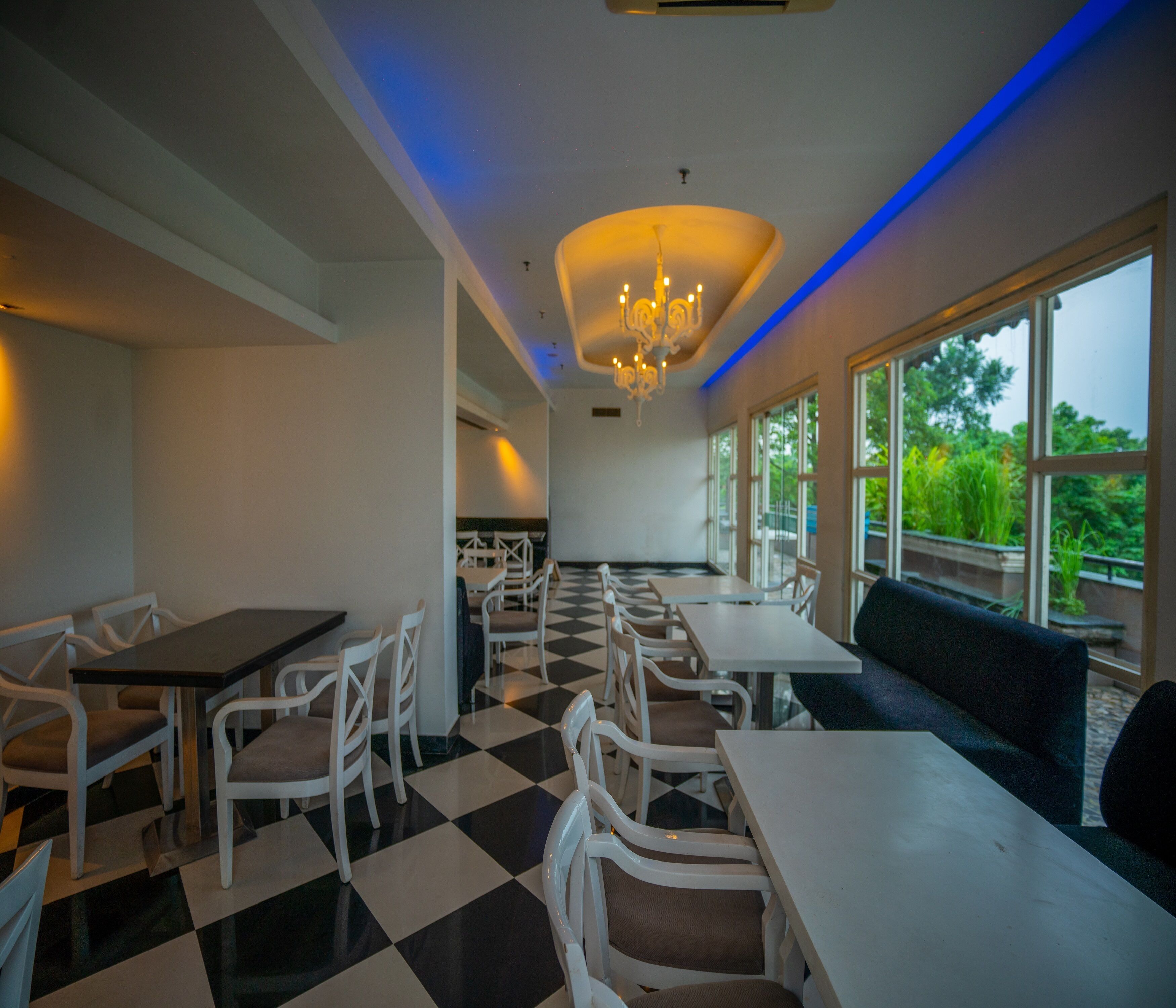 A restaurant with a checkered floor, white tables and chairs, a chandelier, and large windows with a view of greenery.