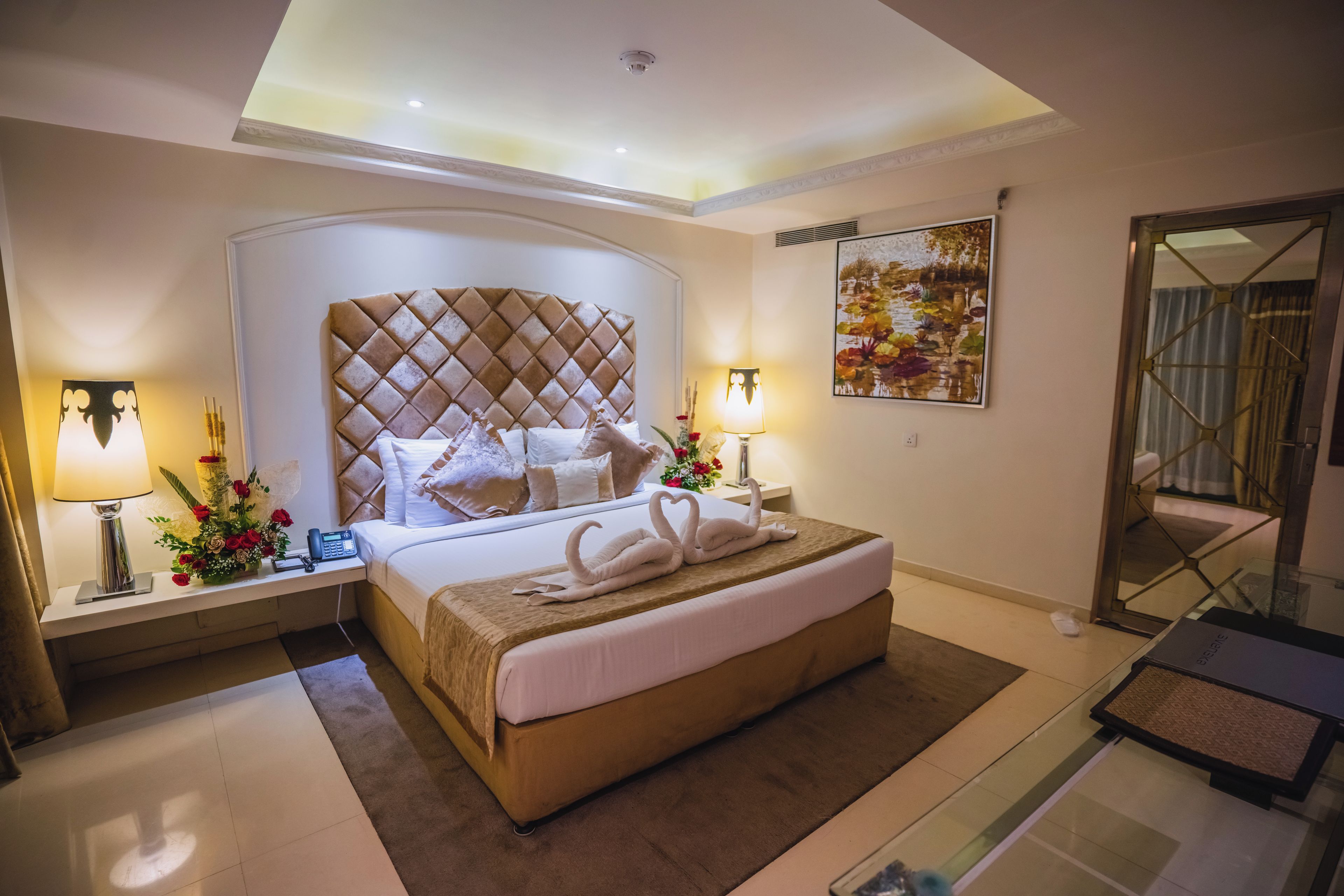 A hotel room with a large bed, two bedside tables with lamps, a decorative wall painting, and a glass partition.