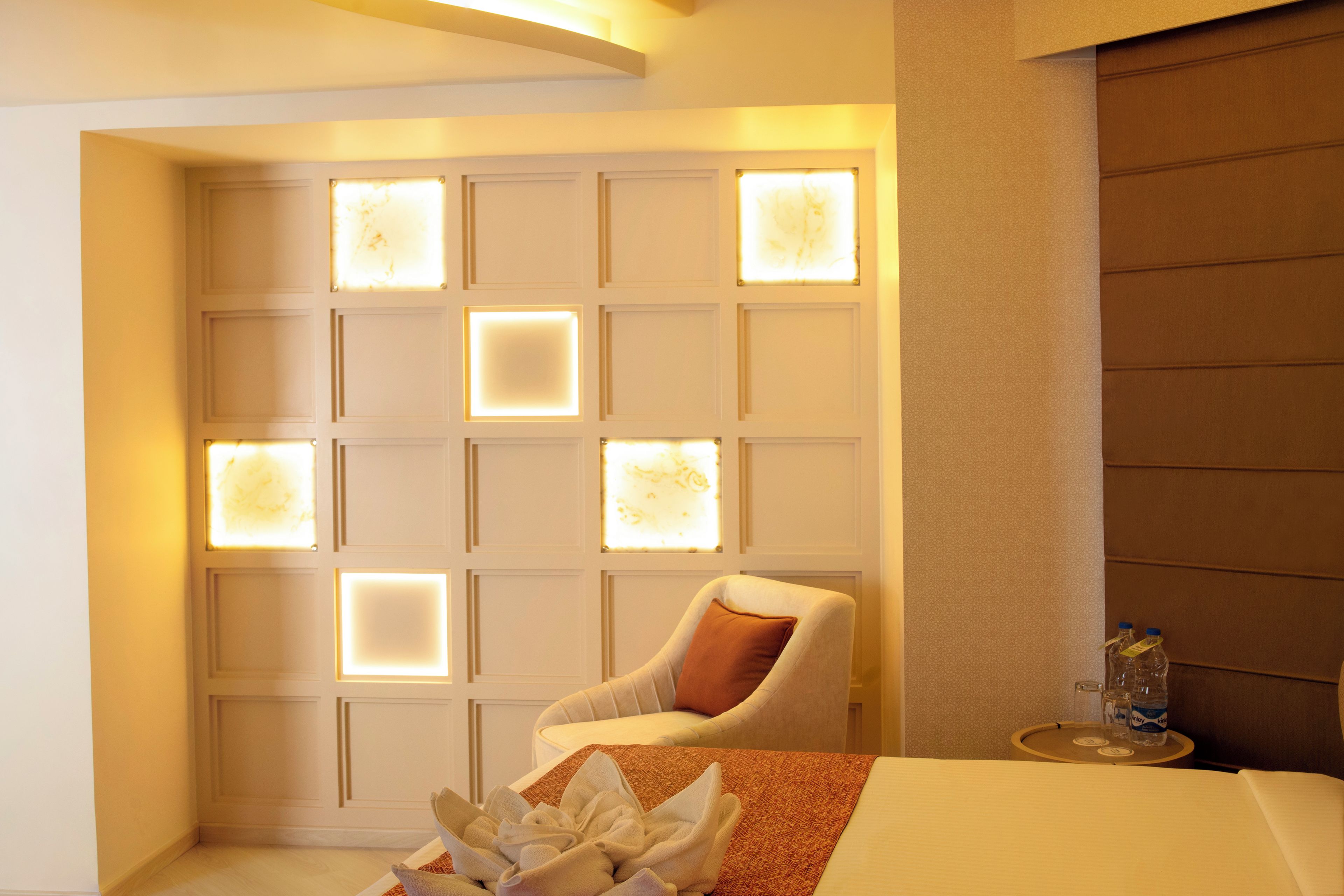 A hotel room with a bed, a chair, a table with water bottles, and a wall with illuminated panels.