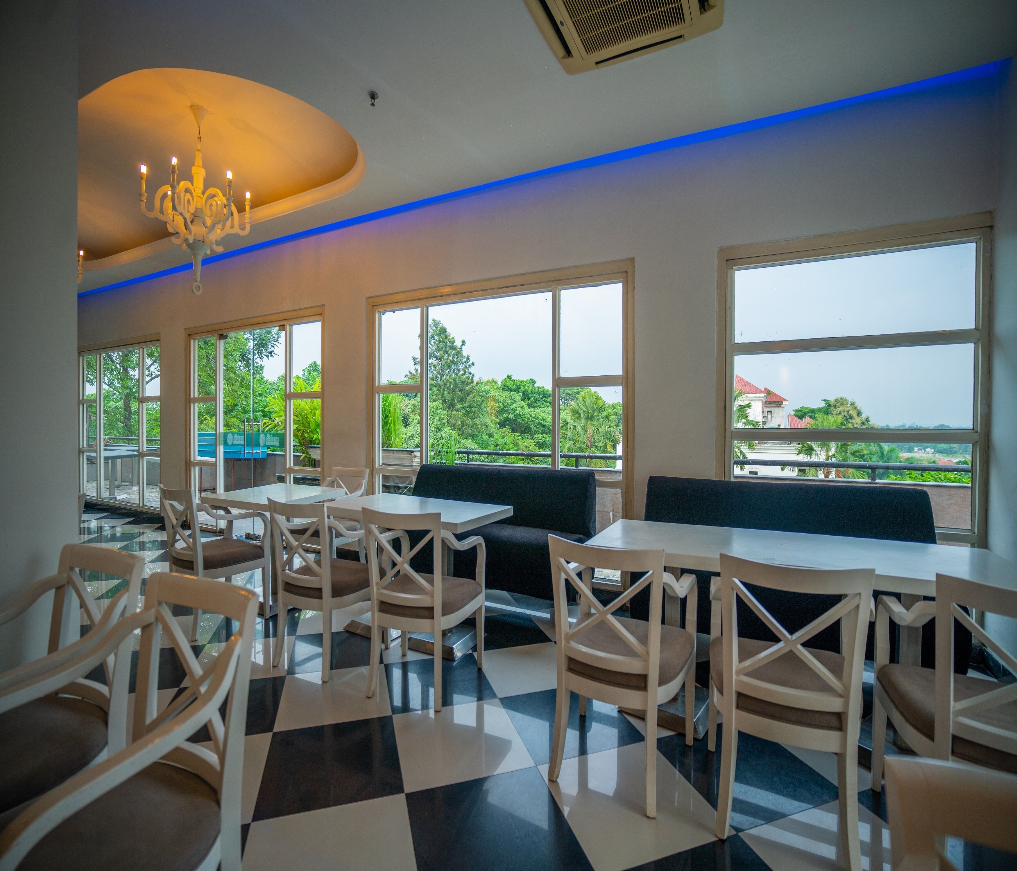 Un restaurant doté d’un sol à carreaux, de tables et de chaises blanches, d’un lustre et de grandes fenêtres offrant une vue sur la verdure et les bâtiments.