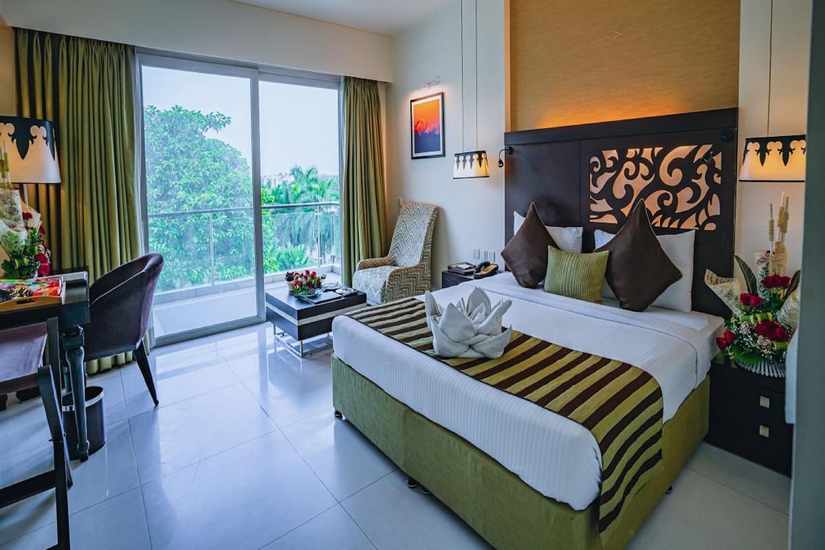 Une chambre d’hôtel avec un grand lit, un bureau et une chaise. Elle dispose d’un balcon avec vue sur la verdure.