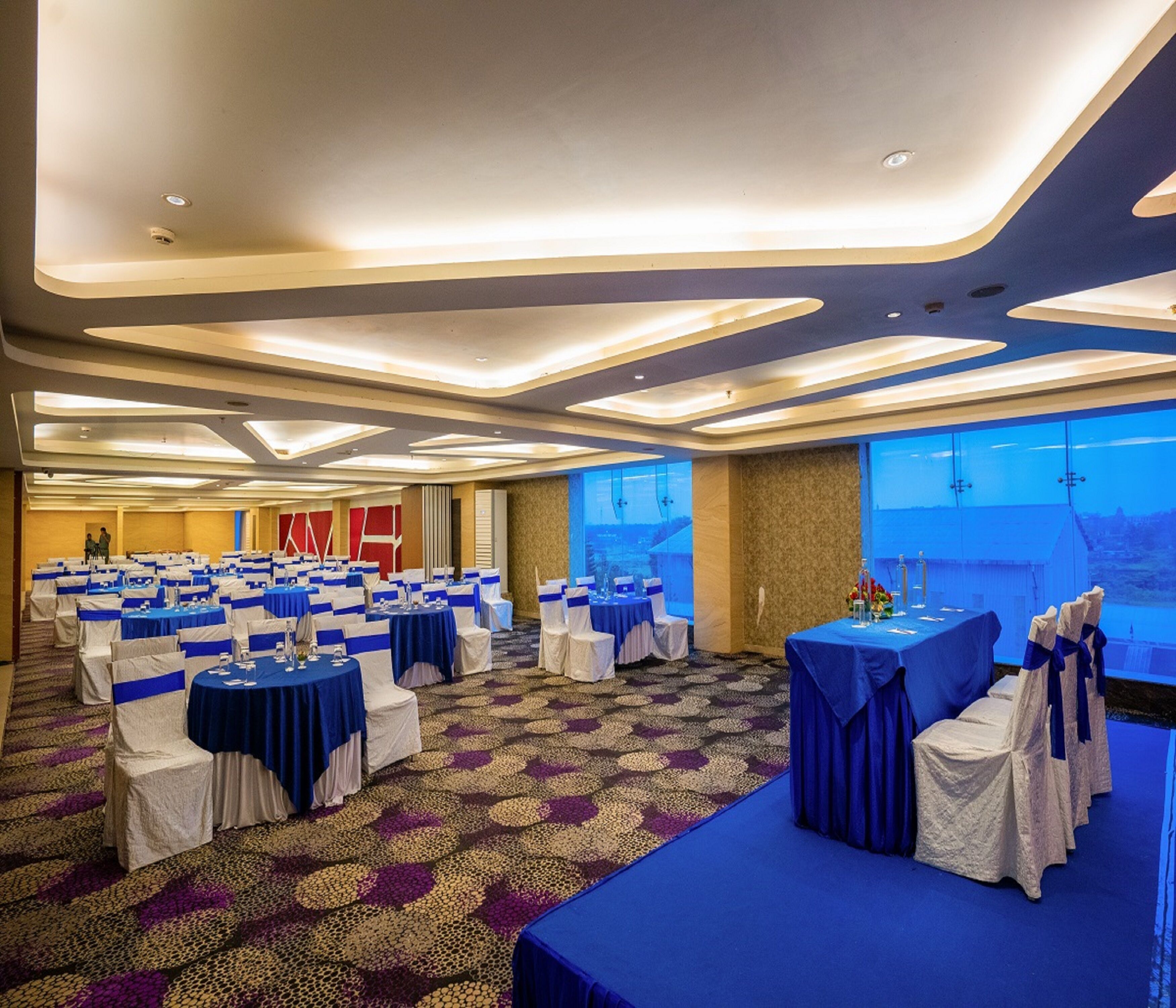 Un salón de conferencias con varias mesas y sillas, un suelo estampado y vistas al exterior a través de amplios ventanales.