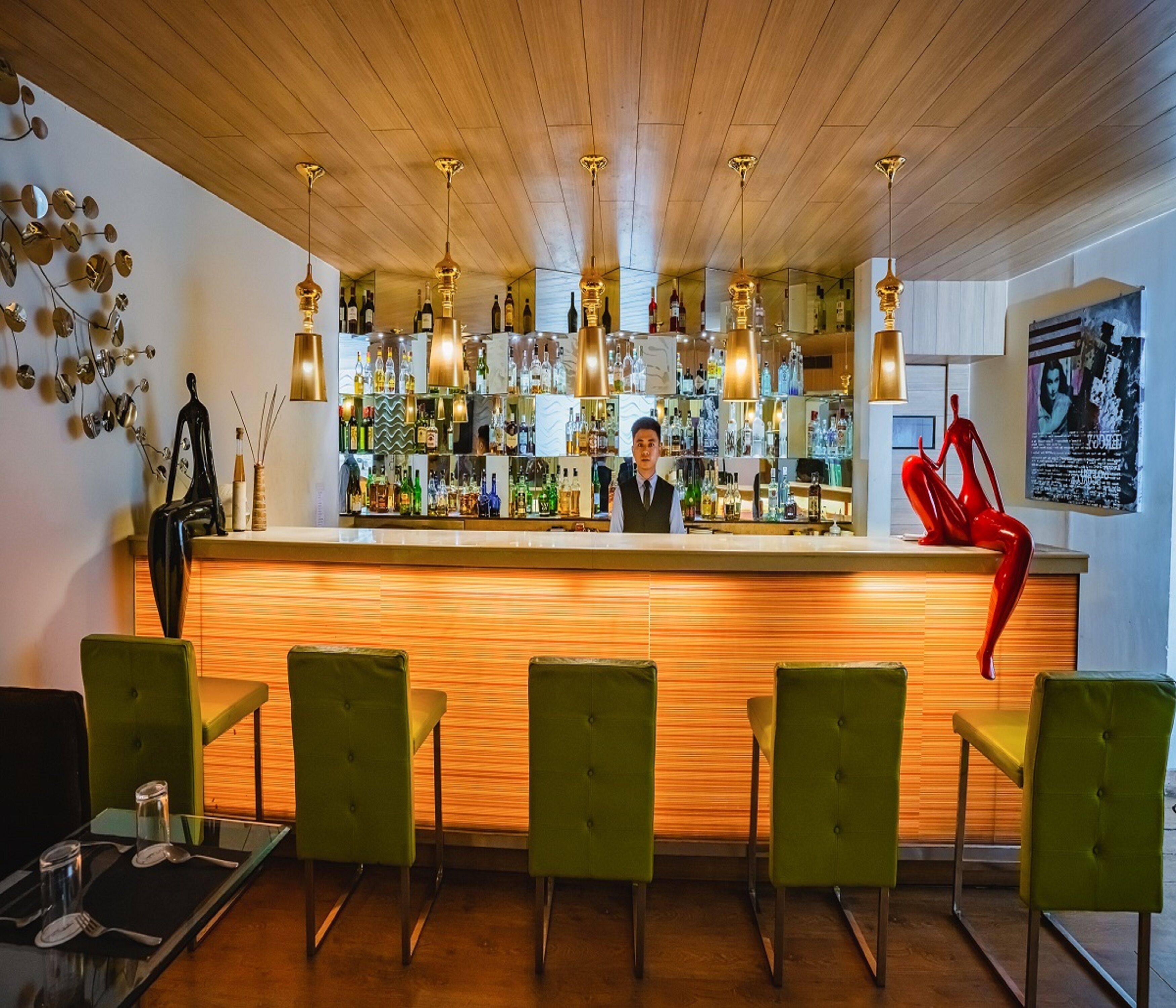 A bar area with a wooden ceiling, green upholstered chairs, and a bar counter with various bottles.