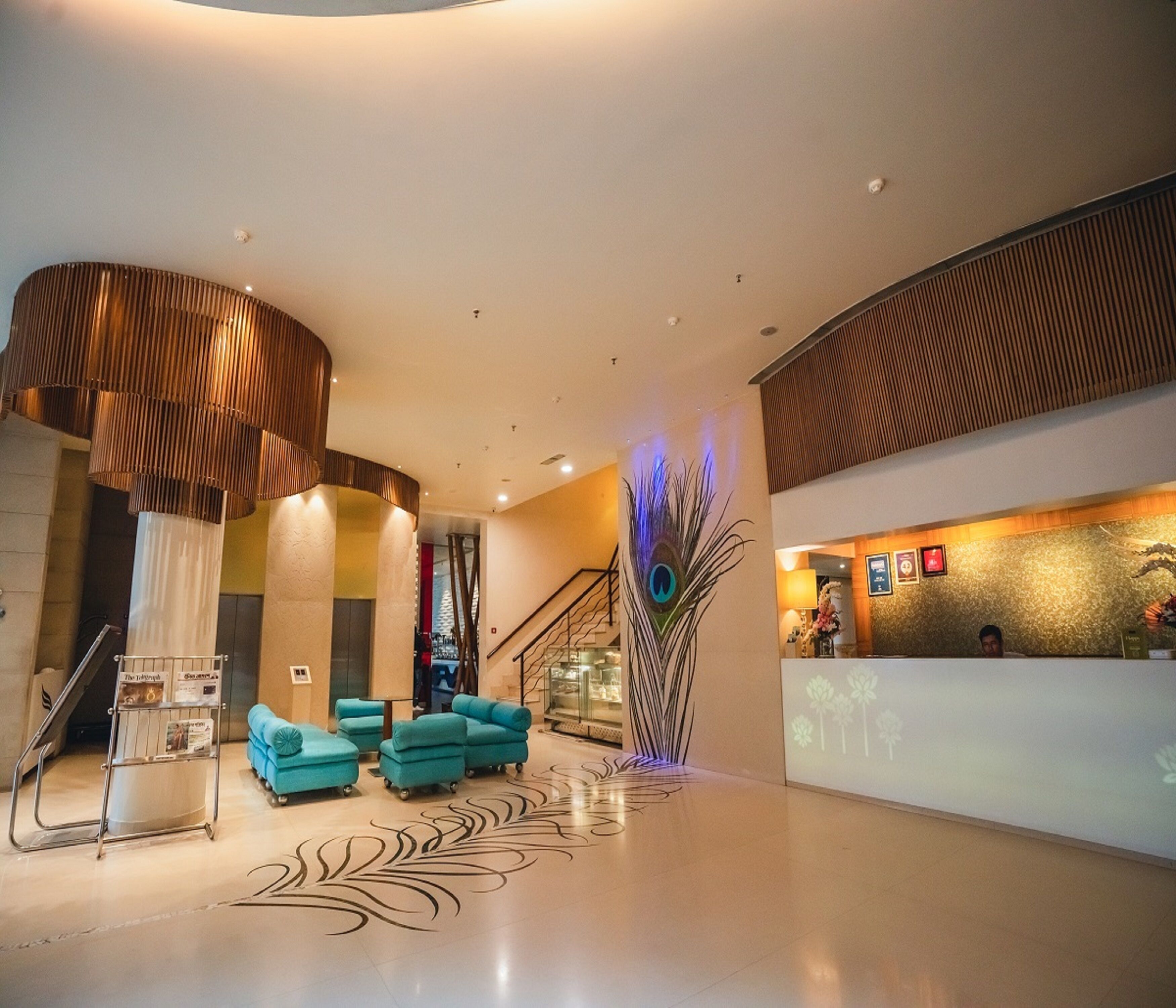 A modern lobby with a reception desk, teal seating, a peacock feather wall art, and a staircase.