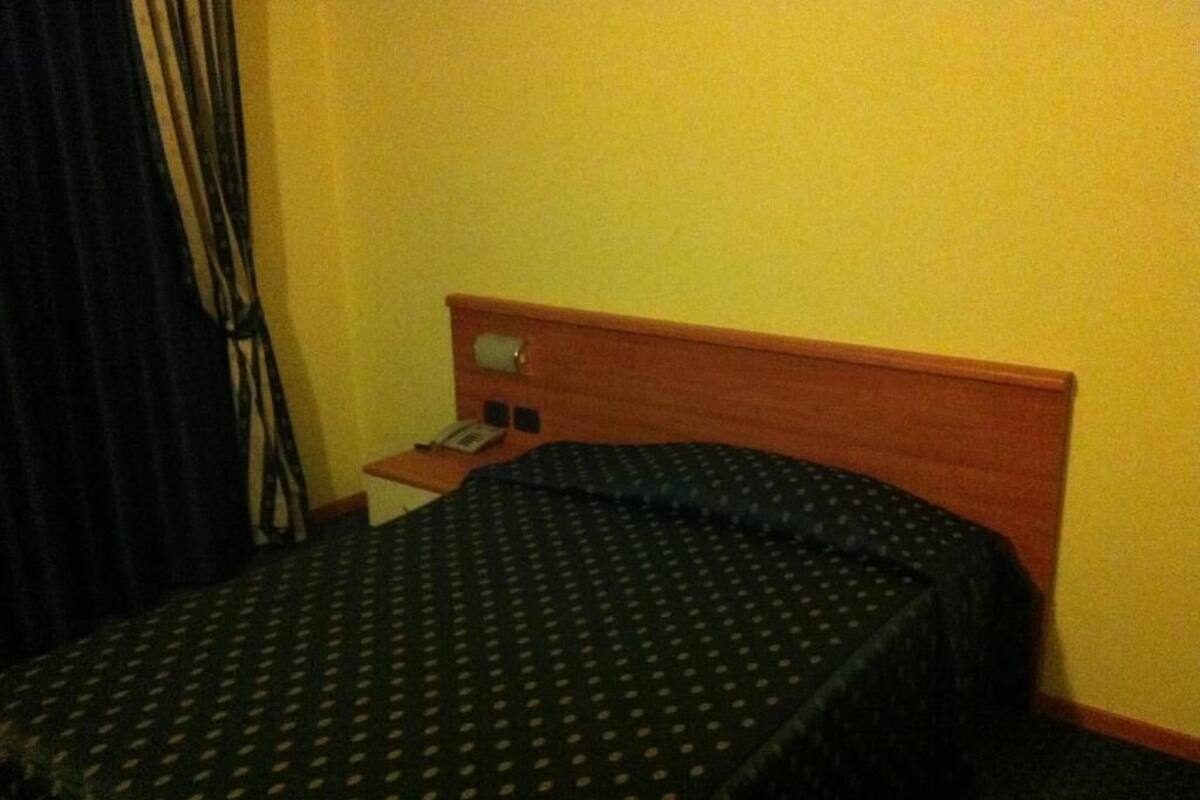 A hotel room with a single bed, a wooden headboard, a nightstand with a telephone, and a curtain.