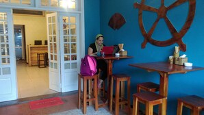 A person seated at a wooden table with a laptop, a pink bag, and a wooden stool. A large wooden wheel decoration is on the wall, and a red mat with "WELCOME" is on the floor. - Like Backpacker Hostel (Da Nang)