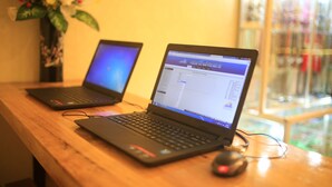 Two laptops on a wooden table with a mouse, a vase with flowers, and a window with a view of a building. - Like Backpacker Hostel (Da Nang)