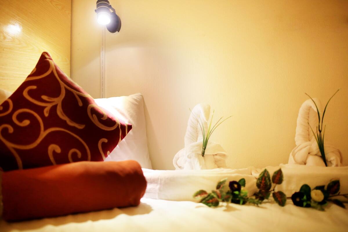 Ein ordentlich bezogenes Bett mit einem roten Kissen mit Muster, weißen Kissen und einem weißen Handtuch. Eine kleine Pflanze und ein Handtuchhalter mit Handtüchern stehen daneben.