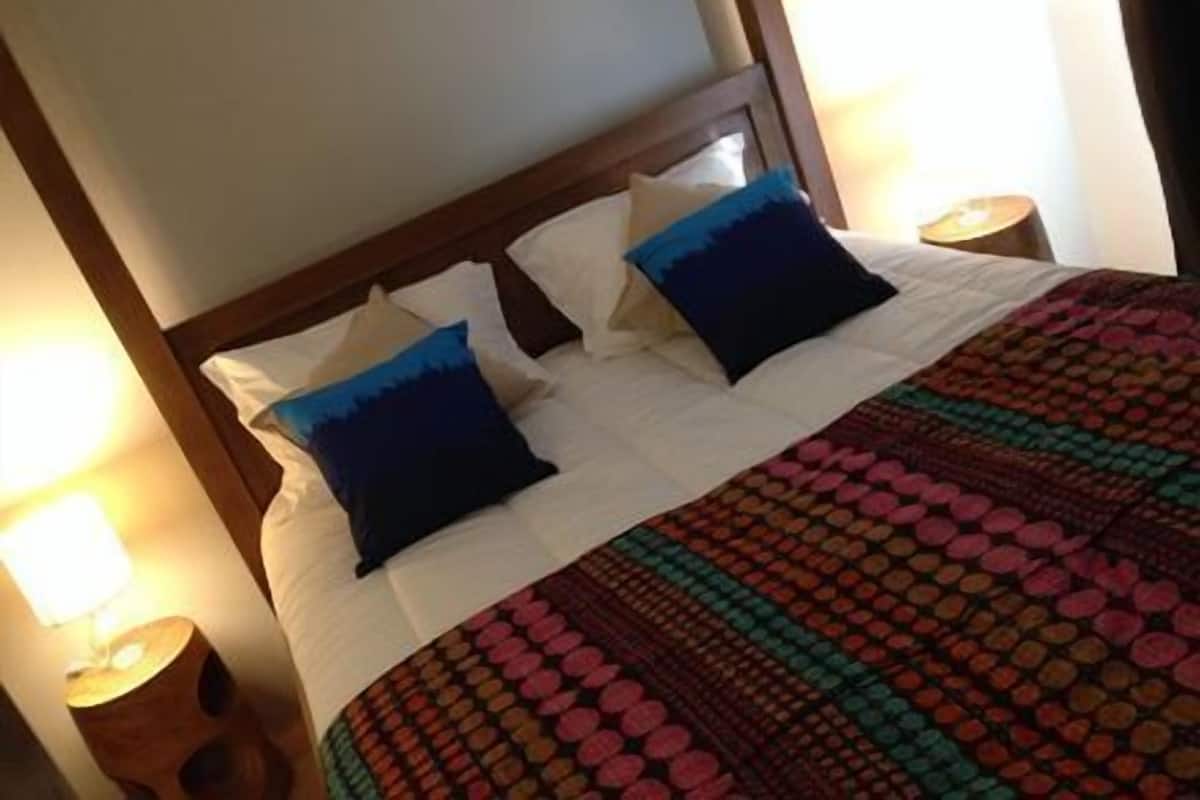 A neatly made bed with white linens, two blue pillows, and a colorful patterned blanket. There are two bedside tables with lamps.