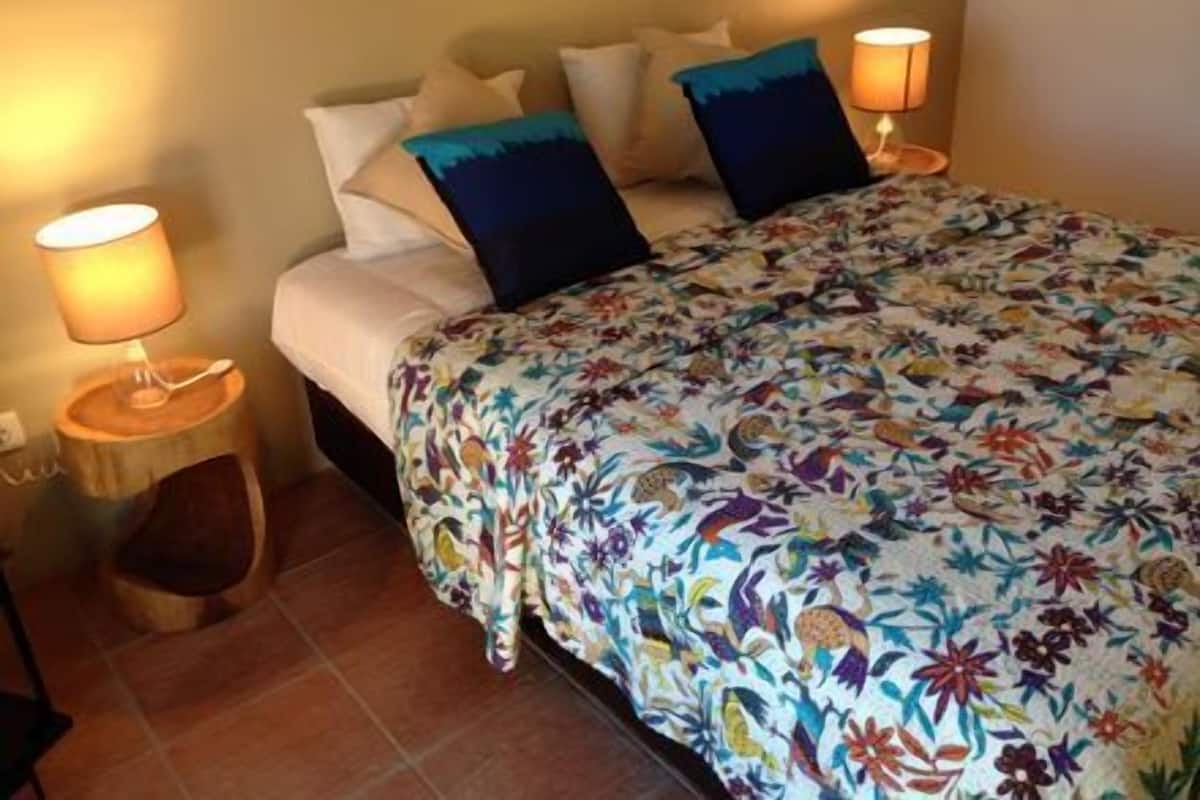 A bedroom with a floral bedspread, two pillows, a nightstand with a lamp, and a wooden chair.