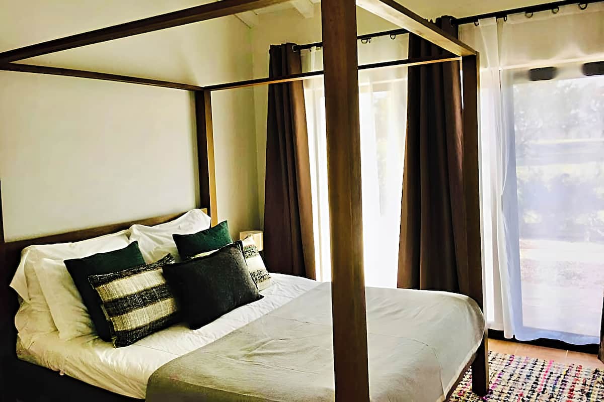 A four-poster bed with a white bedspread and multiple pillows, a wooden headboard, and a view of a garden through sheer curtains.