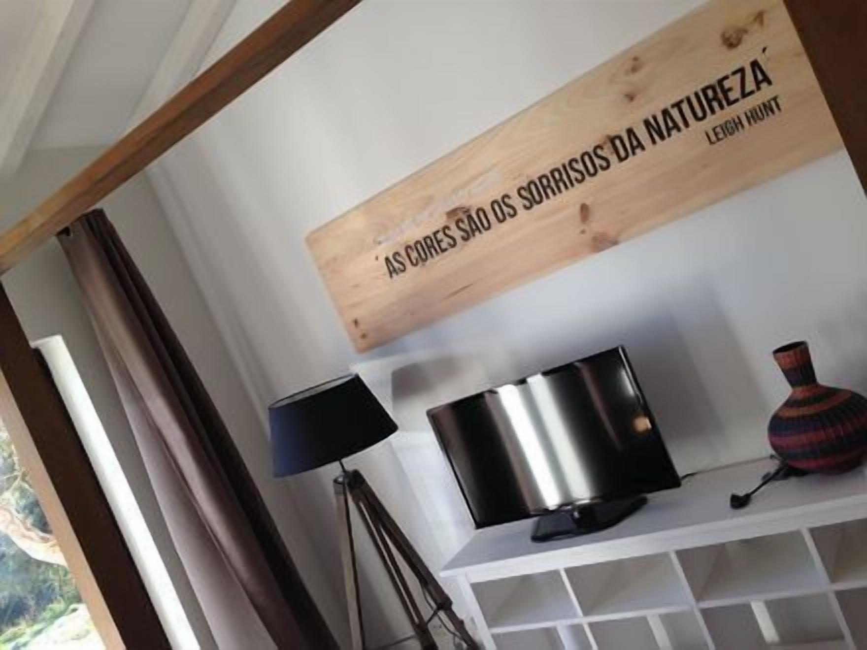 Una habitación con pared blanca, un letrero de madera con texto, una lámpara negra, un objeto metálico y un florero estampado.