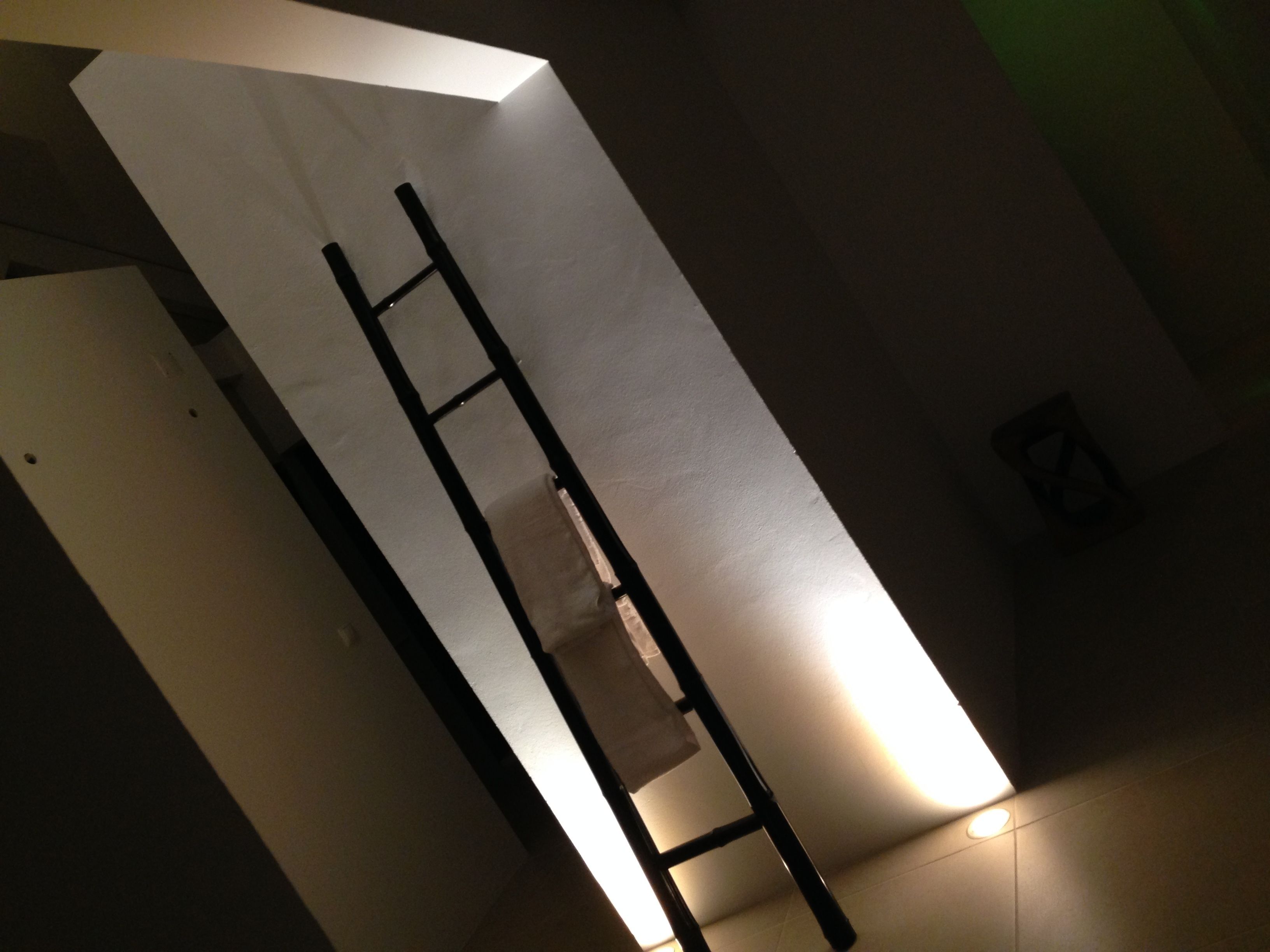 A room with a white wall, a black ladder, and a small light fixture on the floor.