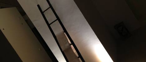 A room with a white wall, a black ladder, and a small light fixture on the floor.