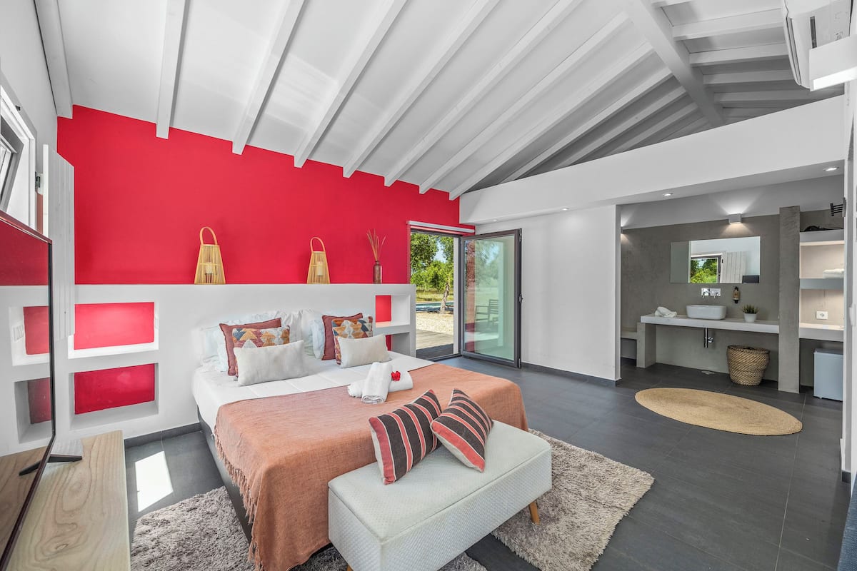 A modern bedroom with a red accent wall, a large bed with multiple pillows, a white bench, and a large window.