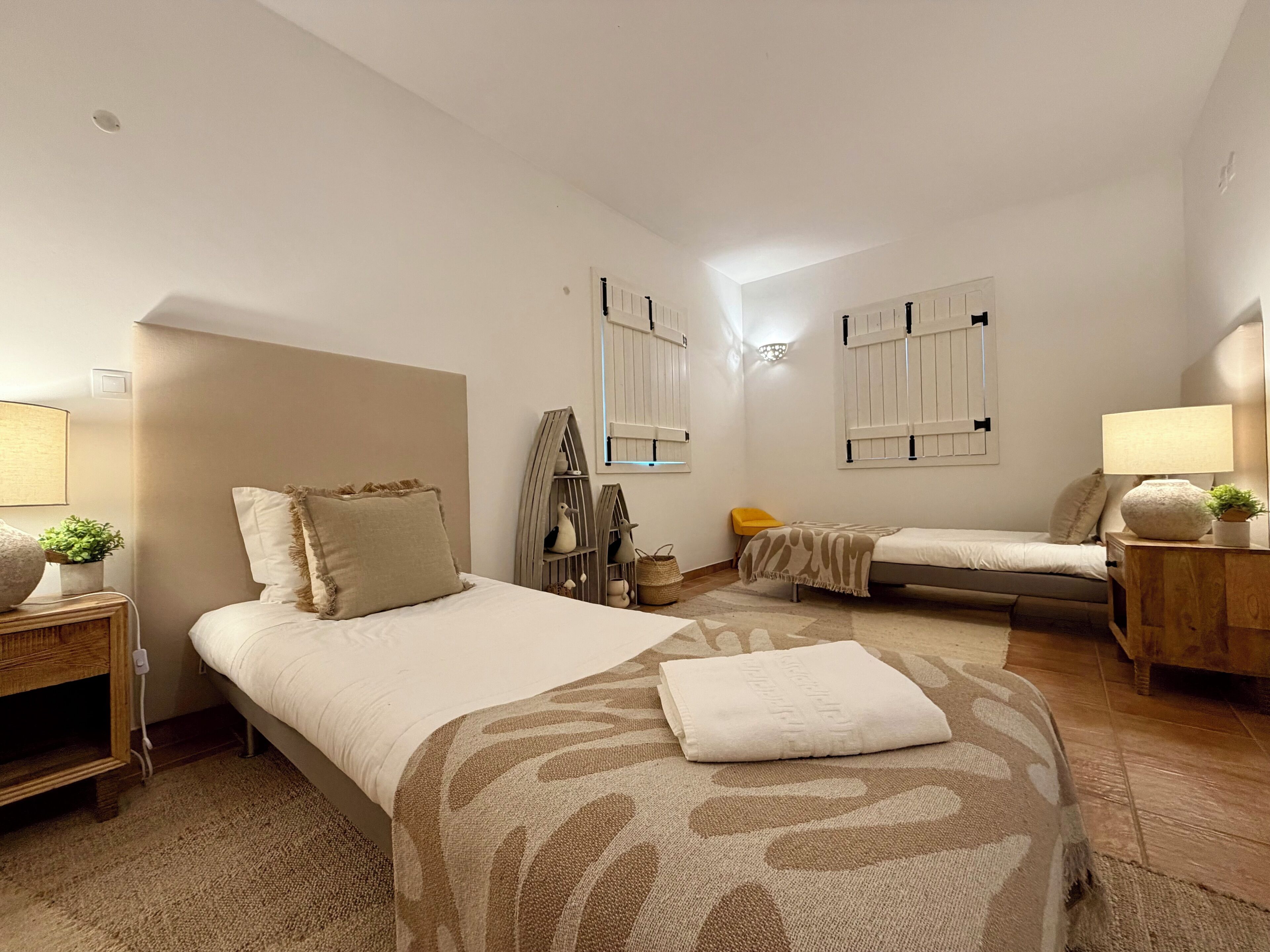 Un dormitorio moderno con una cama grande, una cama más pequeña y un sofá. Hay dos mesitas de noche con lámparas, un estante en la pared y una ventana con contraventanas.