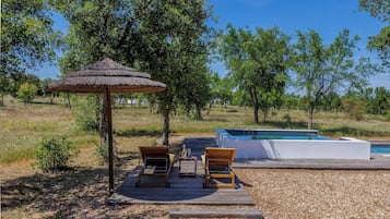 A wooden deck with two lounge chairs and a thatched umbrella, a hot tub, and a pool in a natural setting.