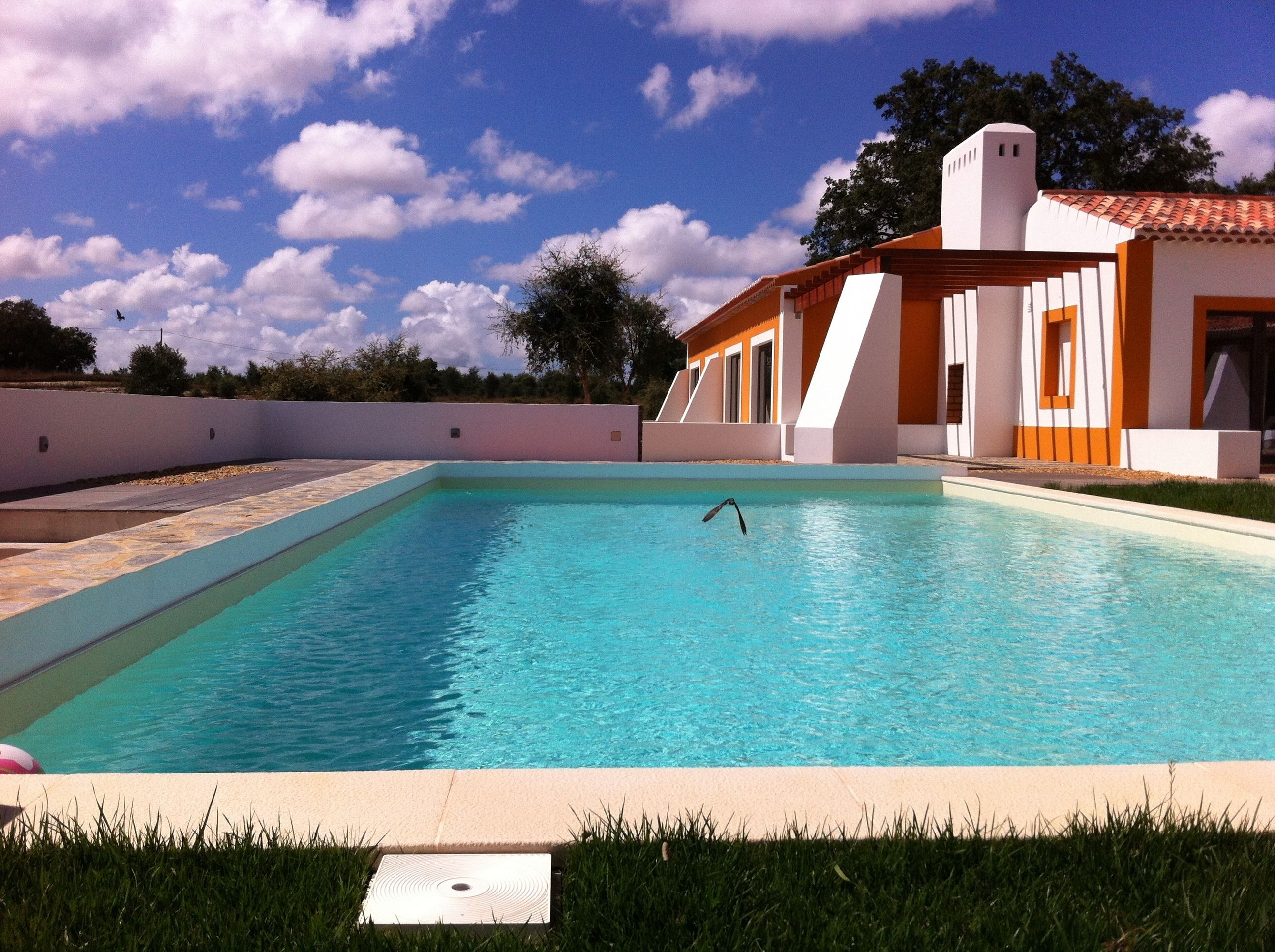 A rectangular swimming pool with clear blue water, surrounded by a white and orange building with a tiled roof, and a grassy area in the foreground.