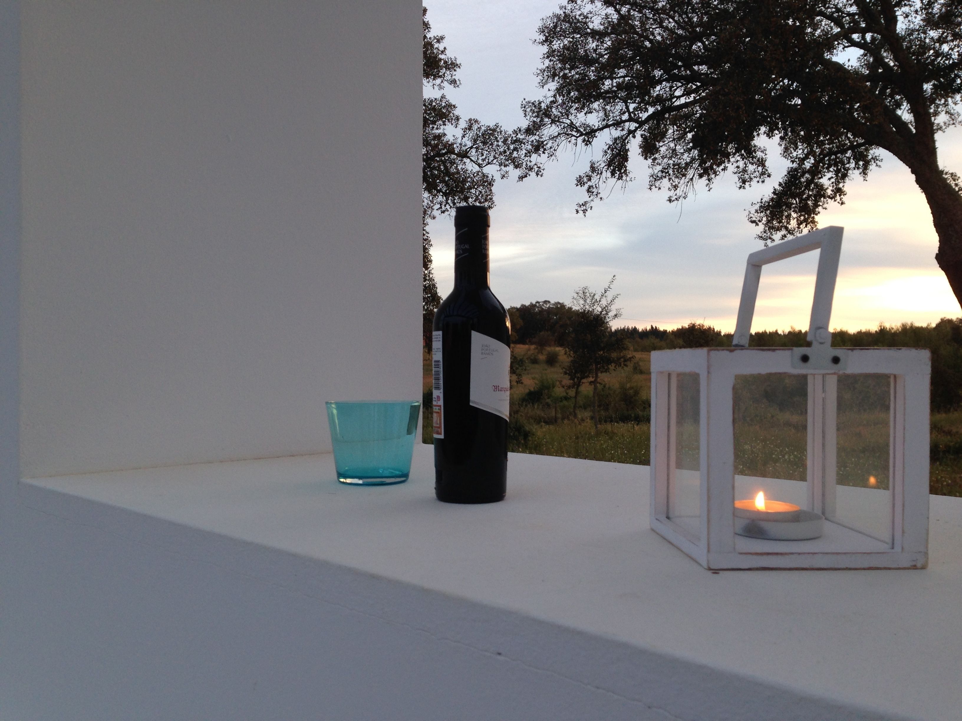 Eine Weinflasche, ein Glas und ein Laternenlicht auf einem Vorsprung mit Blick auf Bäume und einen Sonnenuntergang.