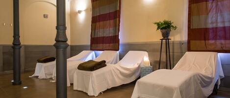 A spa room with two massage tables covered in white sheets, a potted plant, and a candle on a side table.