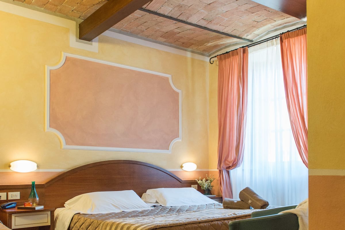 Habitación de hotel con techo de madera, una cama grande con cubrecama acolchado, una mesita de noche con lámpara y una silla con una mesita.