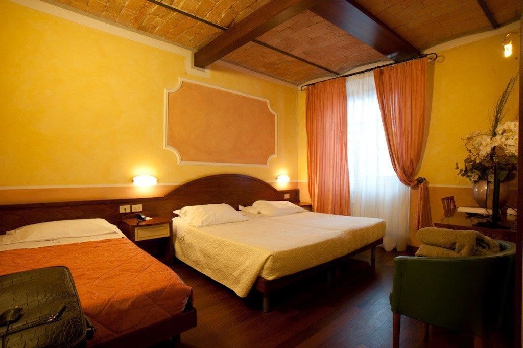 Habitación de hotel con dos camas, cabeceros de madera, una mesita de noche con lámpara, una silla y un florero con flores.