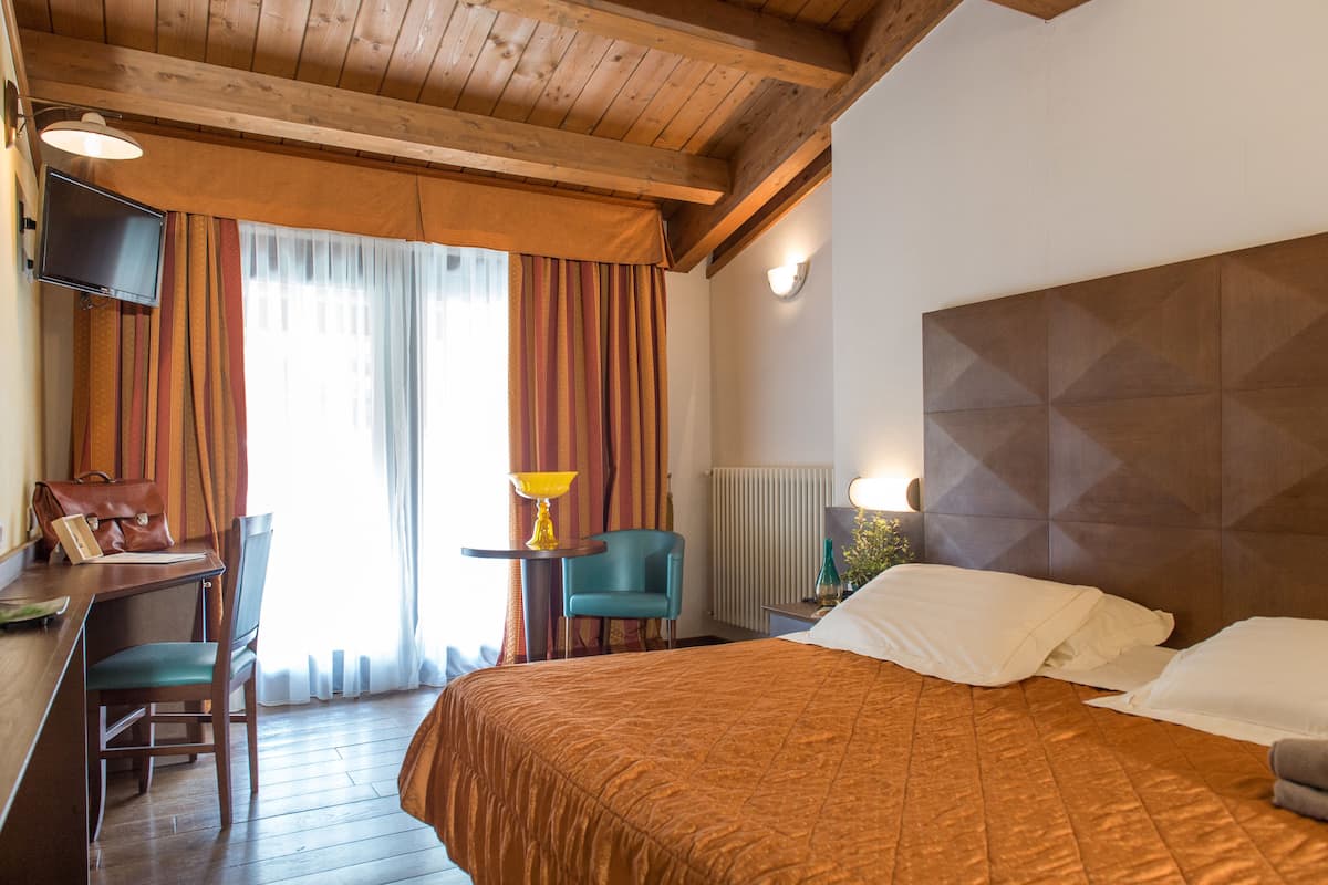 Habitación de hotel con techo de madera, cama con colcha marrón, escritorio con silla, televisor y ventana con cortinas.