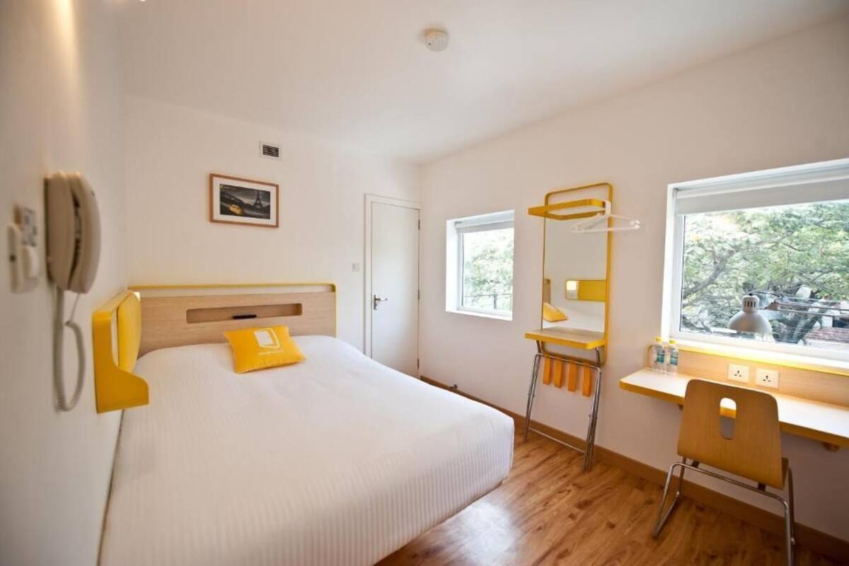 A hotel room with a bed, a desk with a chair, a window with a view, and a mounted phone.