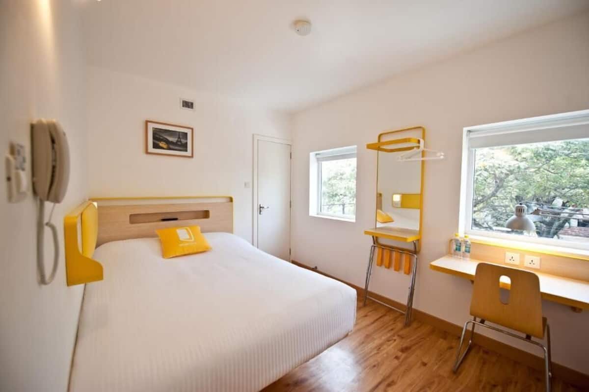 A hotel room with a bed, a desk with a chair, a window with a view, and a mounted phone.