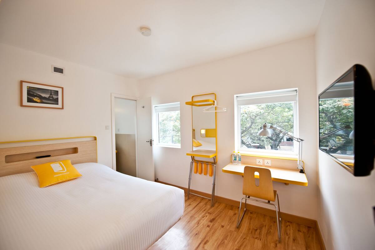 A hotel room with a bed, a desk with a chair, a window, and a picture on the wall.
