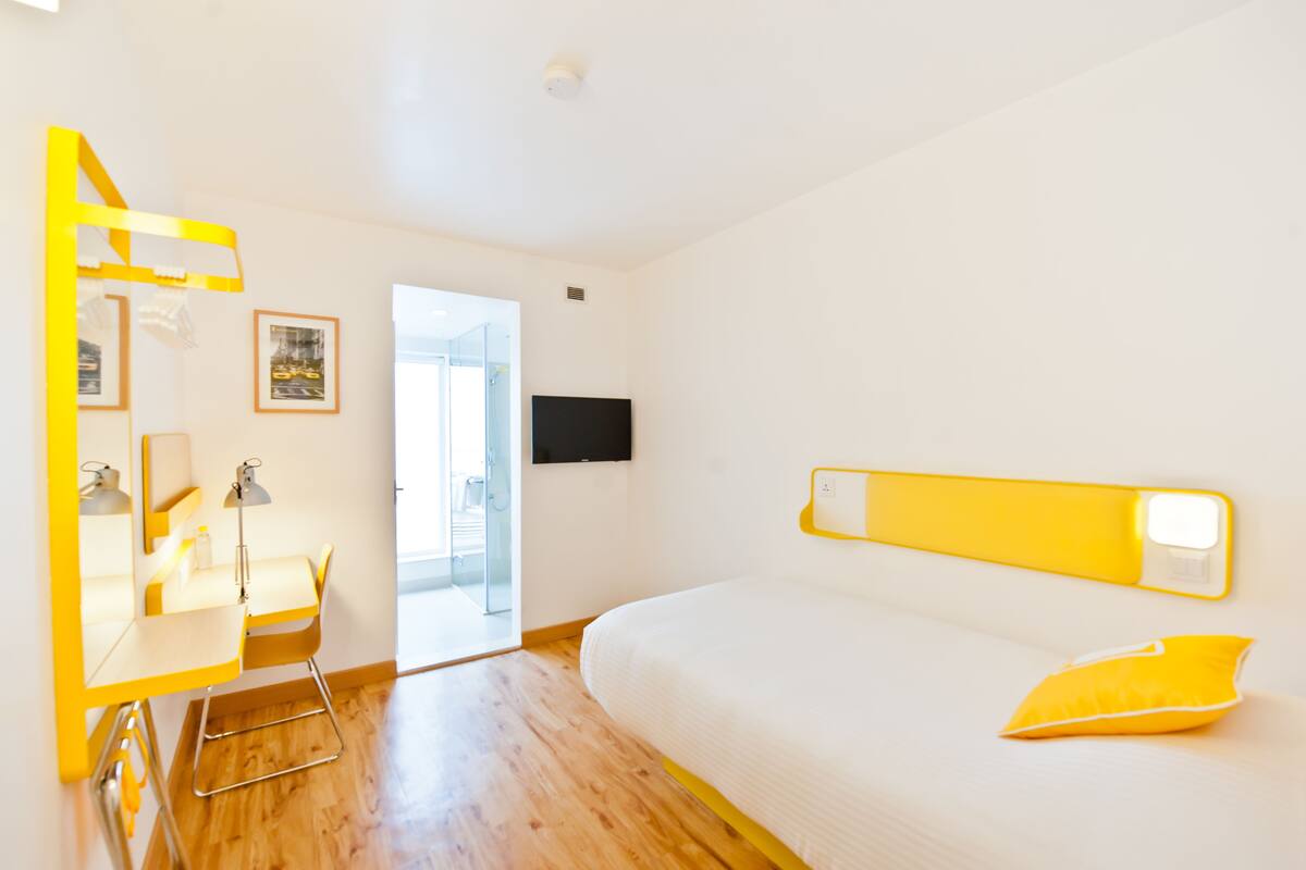 A modern hotel room with a wooden floor, a bed with a yellow pillow, a desk with a chair, a mirror, and a flat-screen TV mounted on the wall.