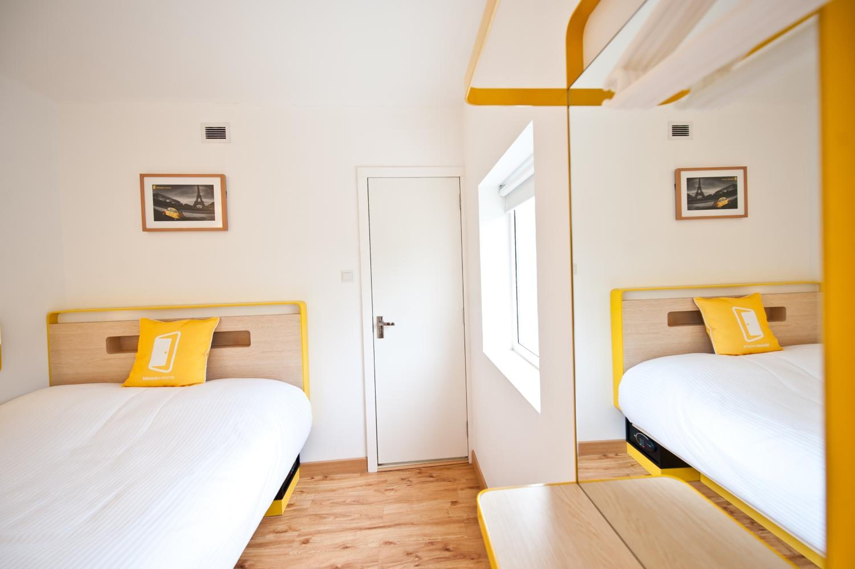 A hotel room with two beds, each with a wooden headboard and a yellow pillow. There are framed pictures on the walls, a door, and a window.