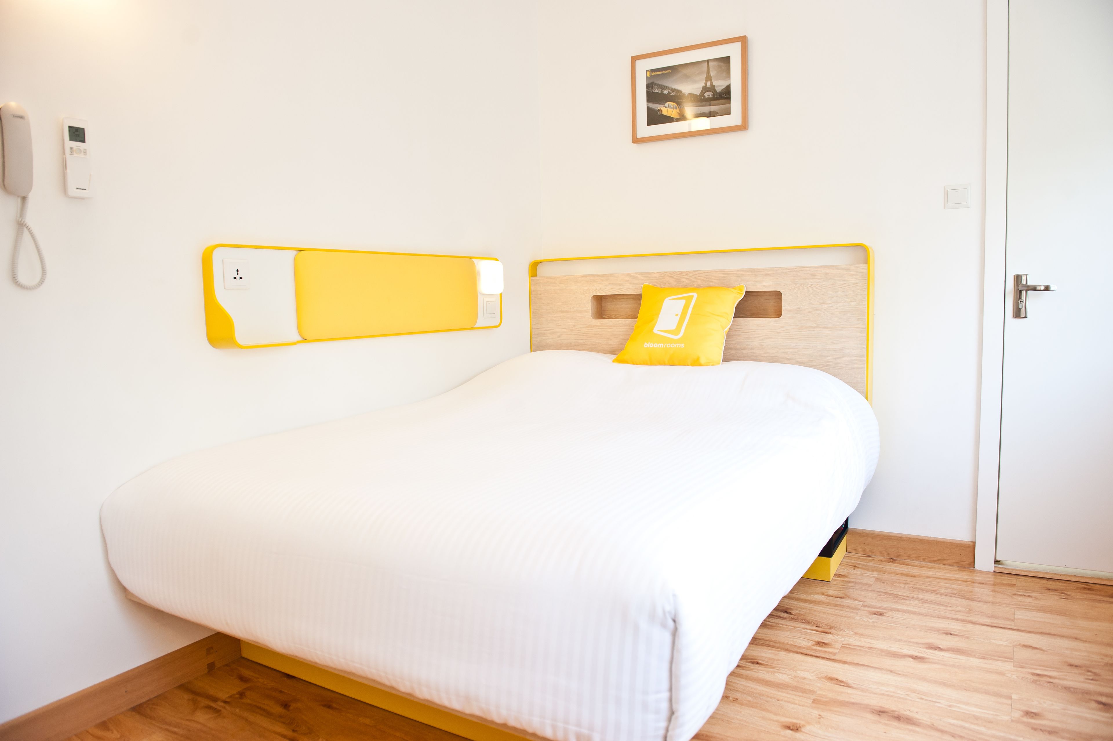 A neatly made bed with a wooden headboard, a bedside table with a yellow lamp, and a wall-mounted phone.