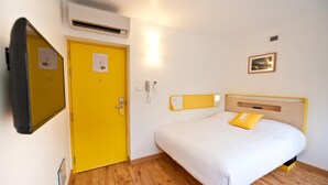 Et hotelværelse med en seng, en gul dør, en aircondition, et natbord, en vægtelefon og et indrammet billede.