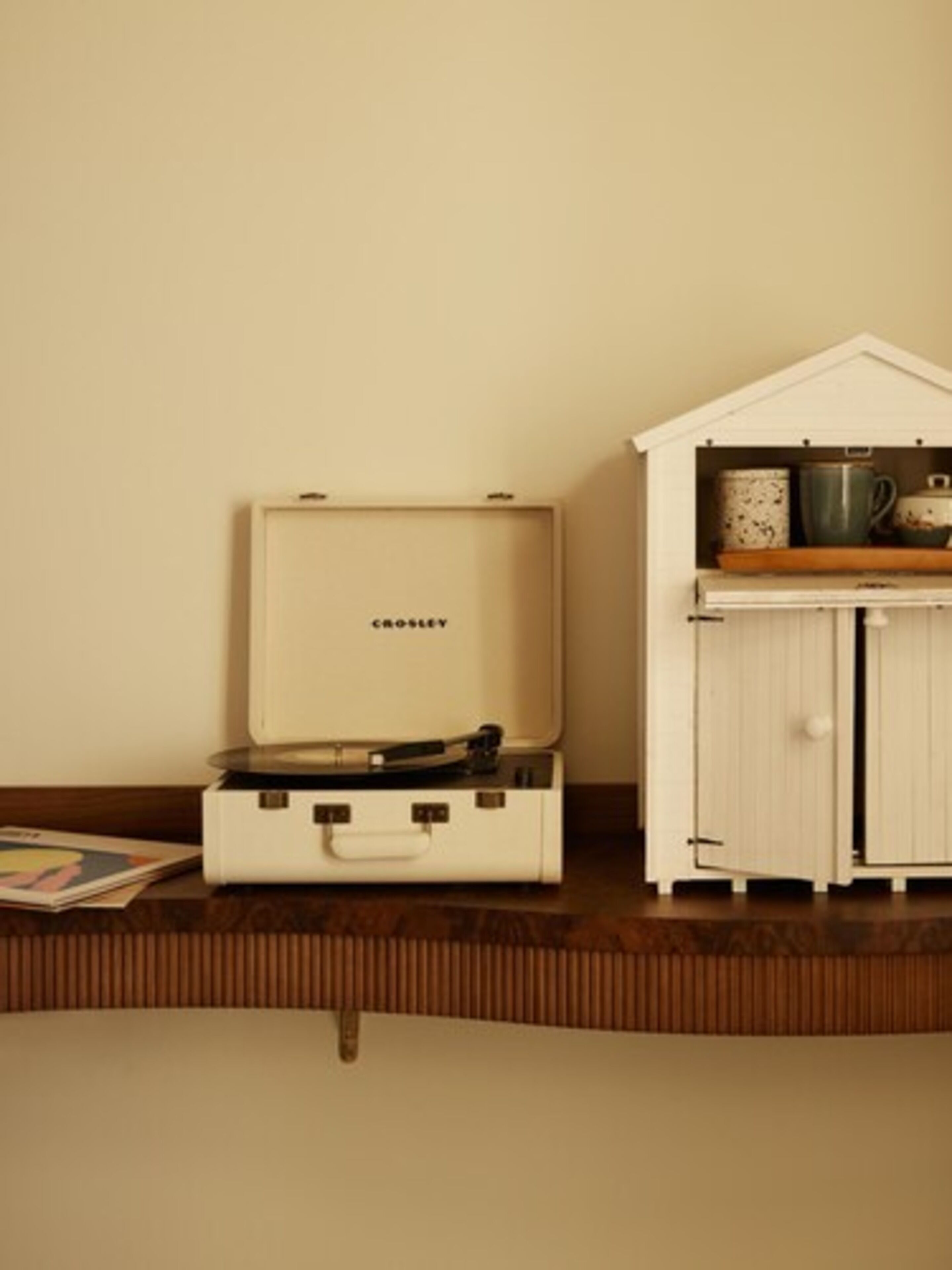 A room with a vintage suitcase, a small white cabinet with kitchenware, and a wooden shelf.