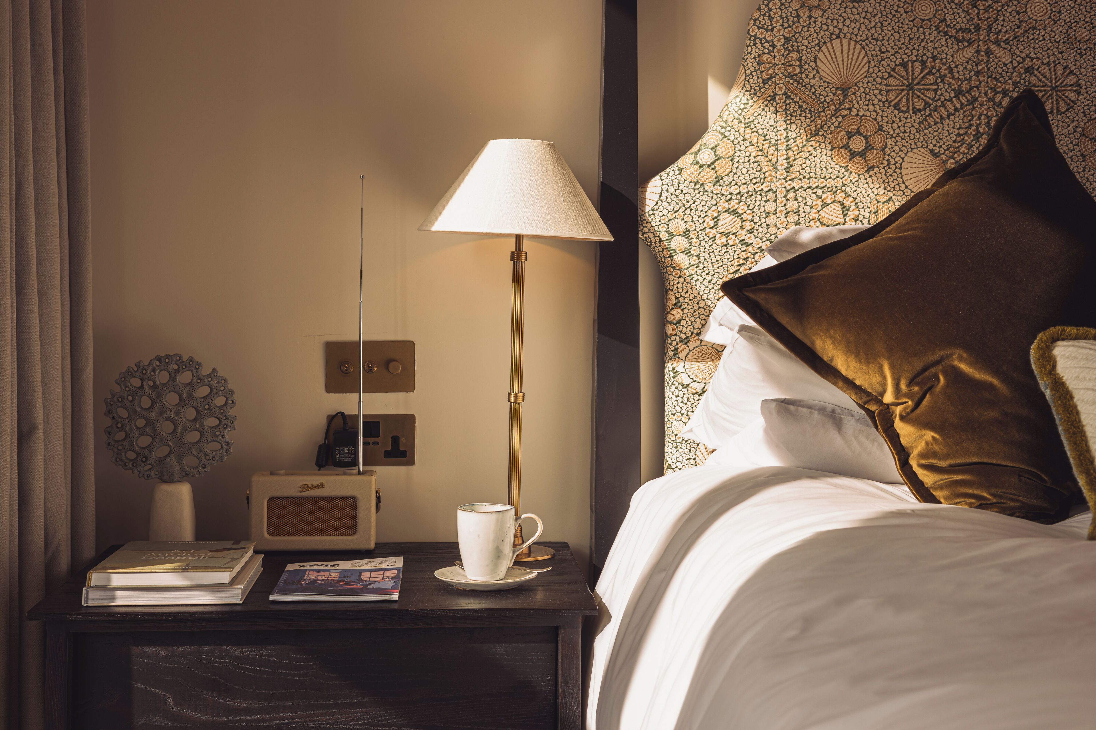 Ein ordentlich bezogenes Bett mit einem braunen Kissen, ein Nachttisch mit einer Lampe, einer Tasse und einem Buch.