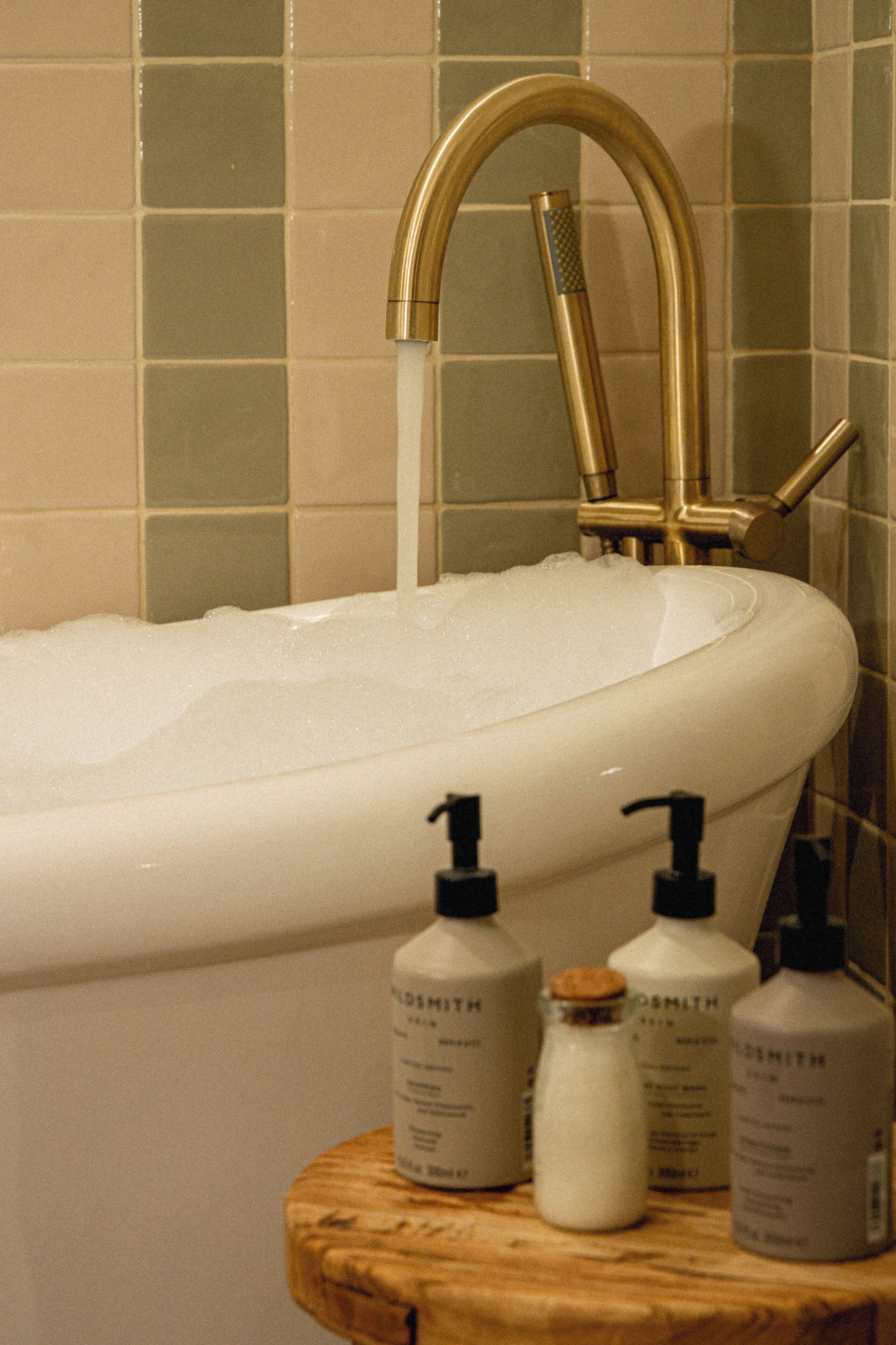 A bathtub with a gold faucet and soap bottles on a wooden stool.