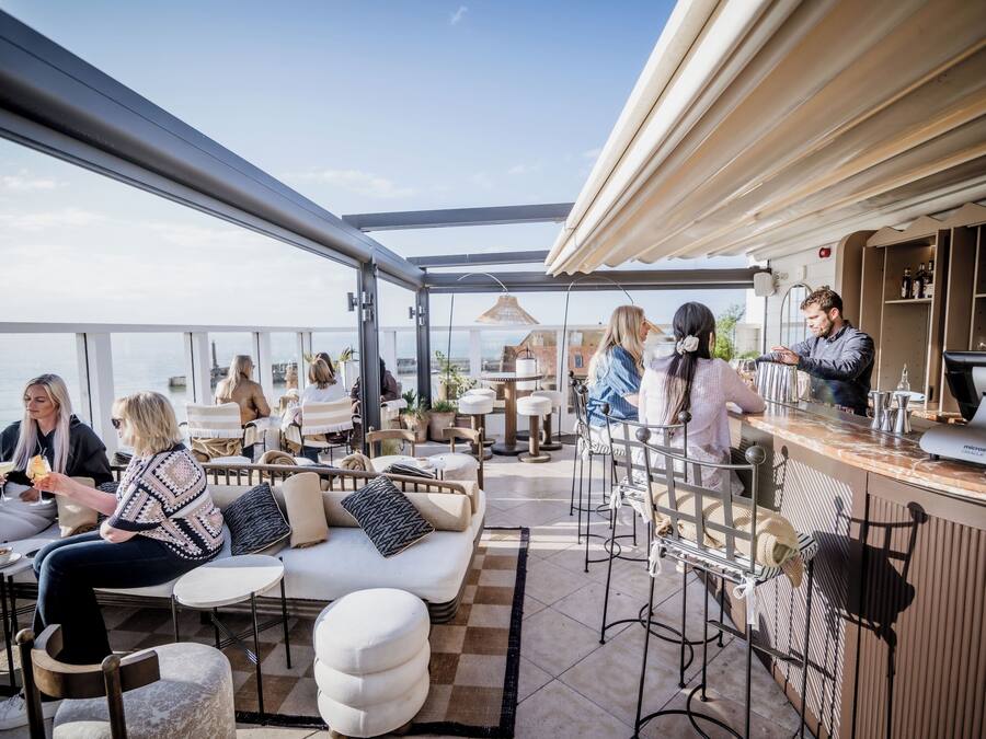 An outdoor seating area with a bar, cushioned seating, and a view of the sea.