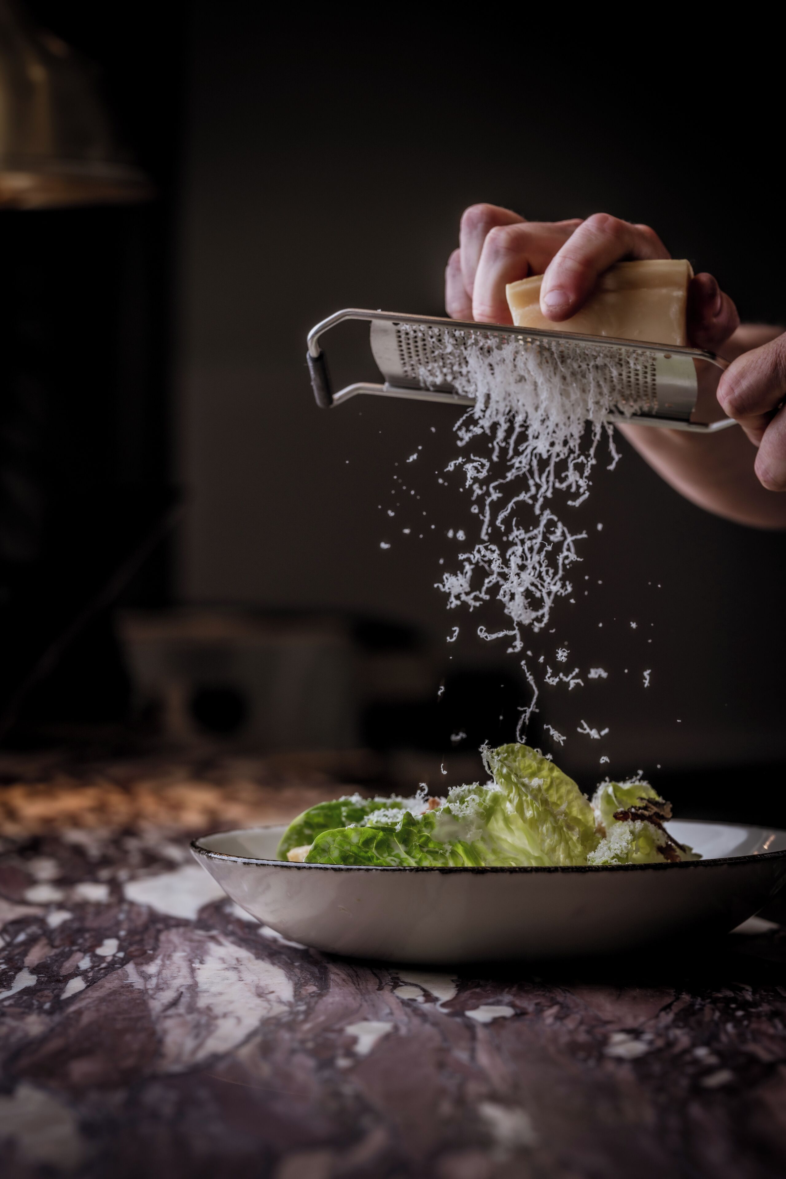 A person grating cheese over a dish of greens.